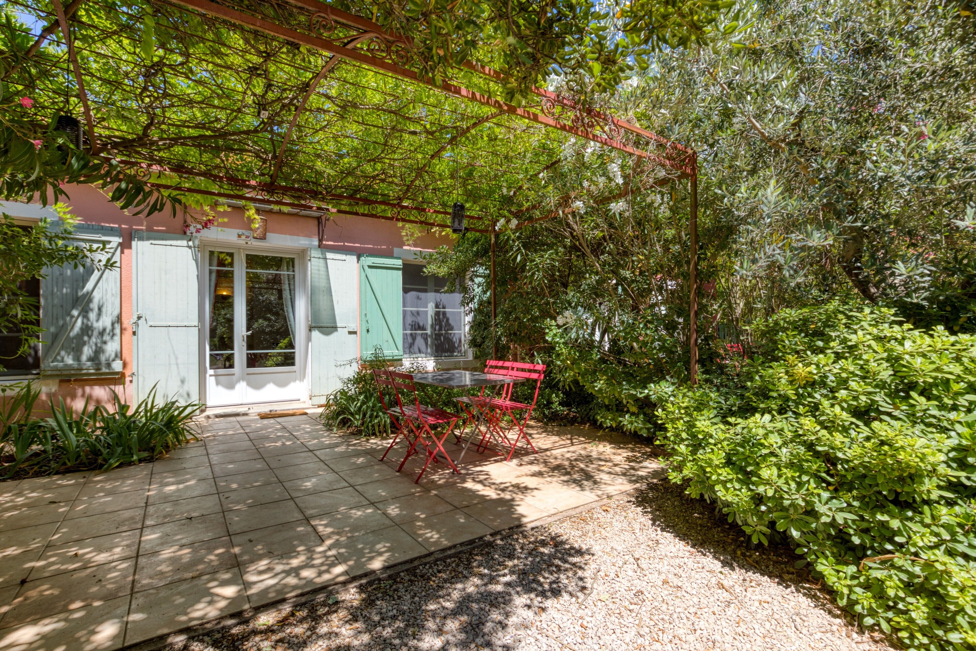 A covered patio with a table and chairs, surrounded by greenery and a building with a door and windows.