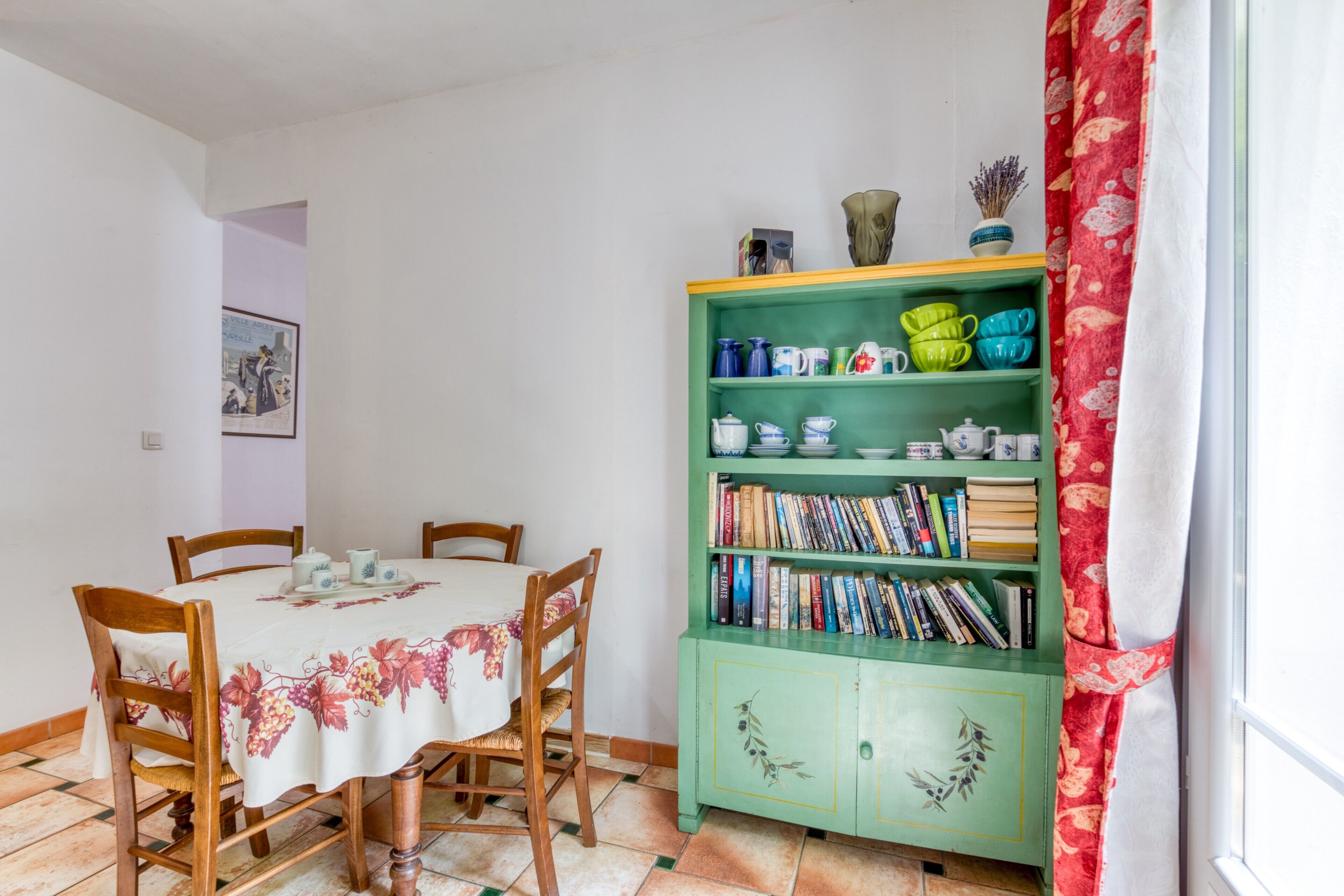A dining area with a table set for four, a green bookshelf with books and decorative items, and a window with a red curtain.