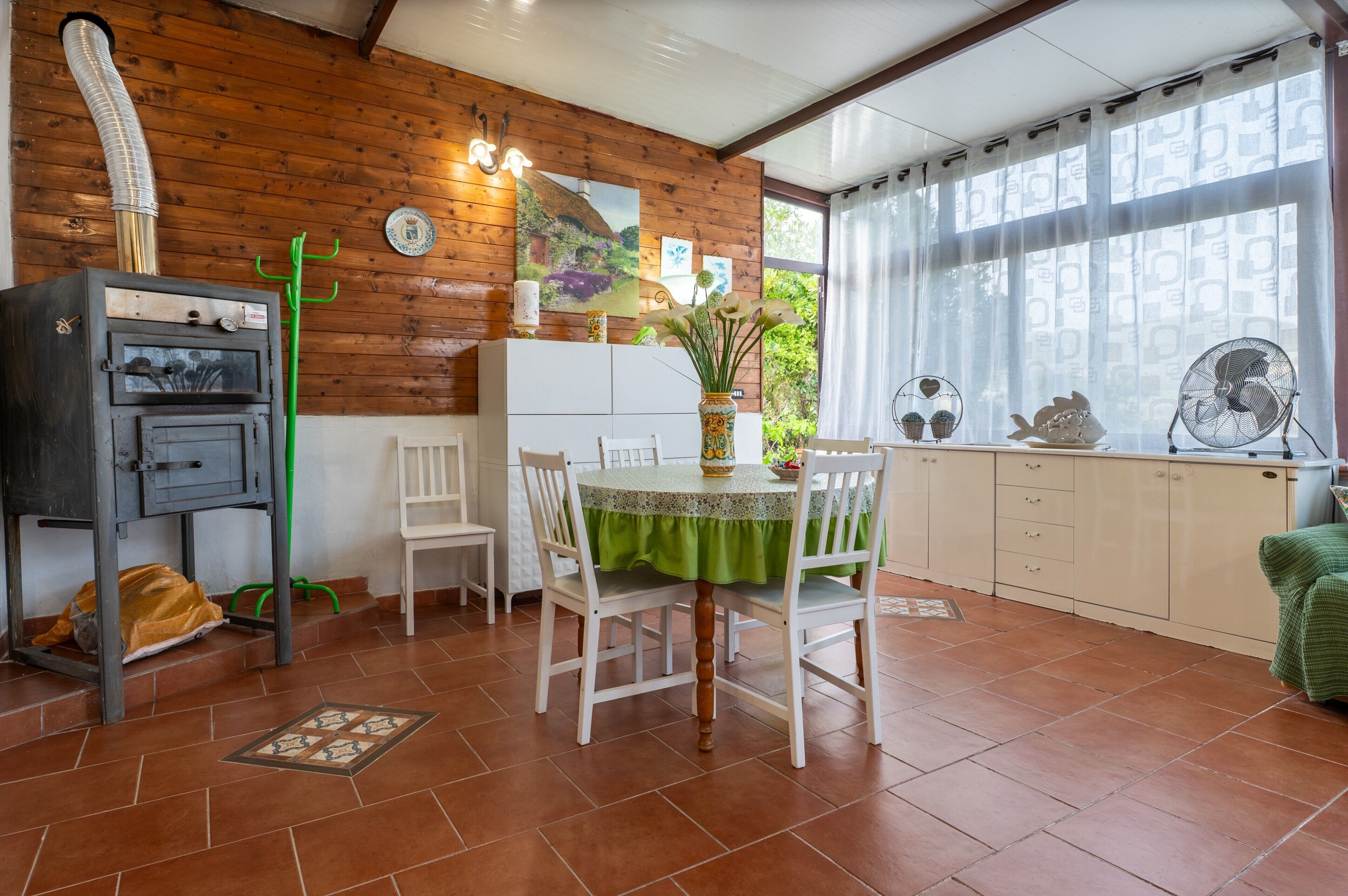 Een keuken met een fornuis, witte kastjes, een eettafel met stoelen en een raam met gordijnen.