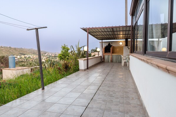 Un balcon offrant une vue sur un paysage vallonné, un sol carrelé et un espace détente couvert équipé d’un barbecue.