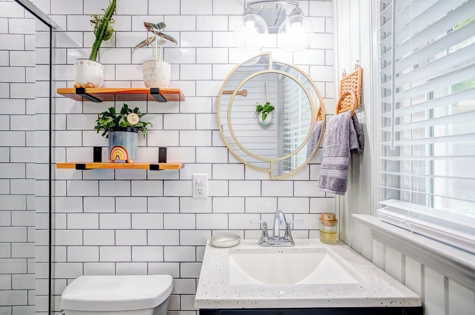 A bathroom with white subway tiles, a white sink, a toilet, wooden shelves with plants, a round mirror, and a window with blinds.