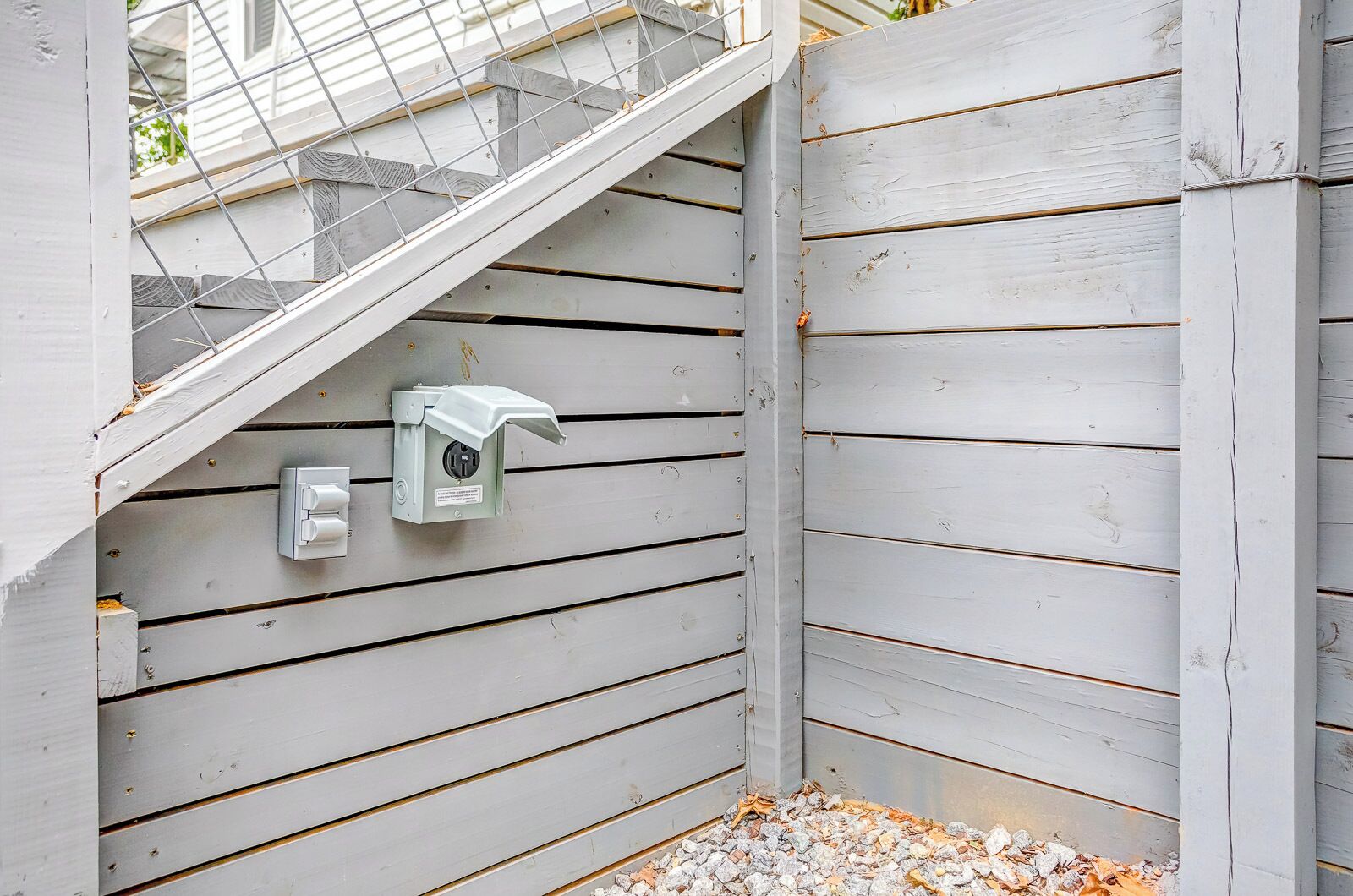 Un angolo di un edificio con rivestimento in legno bianco, una scala in metallo e un quadro elettrico con una presa.