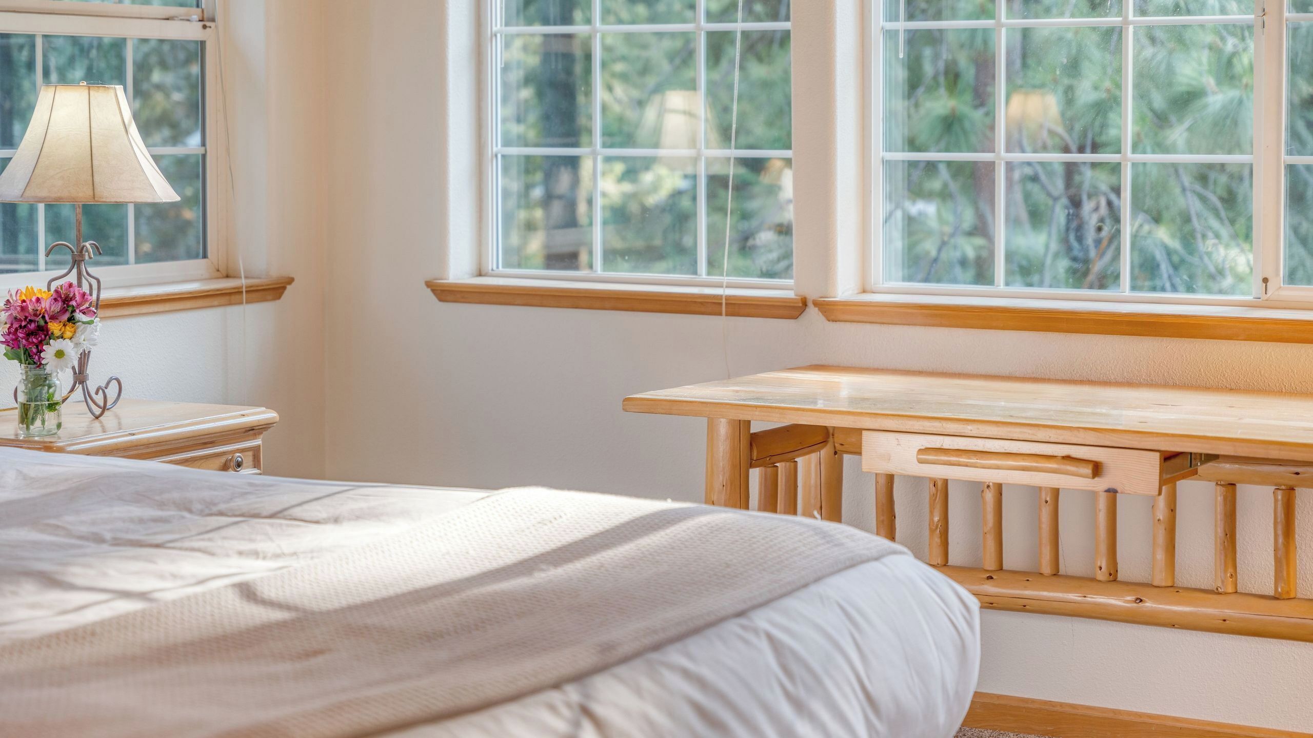 A bedroom with a bed, a wooden bench, a nightstand, a lamp, and a window with a view of greenery.