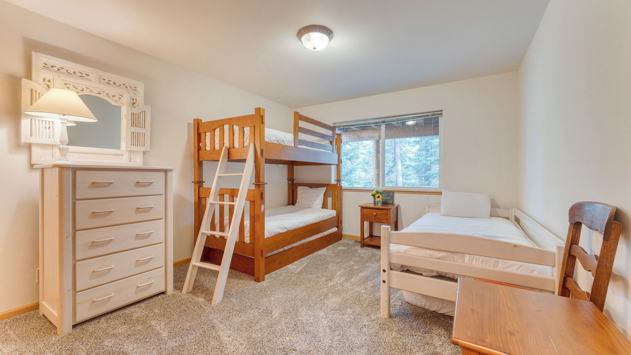 Une chambre avec des lits superposés, une commode, un miroir, une fenêtre donnant sur les arbres et une lampe.