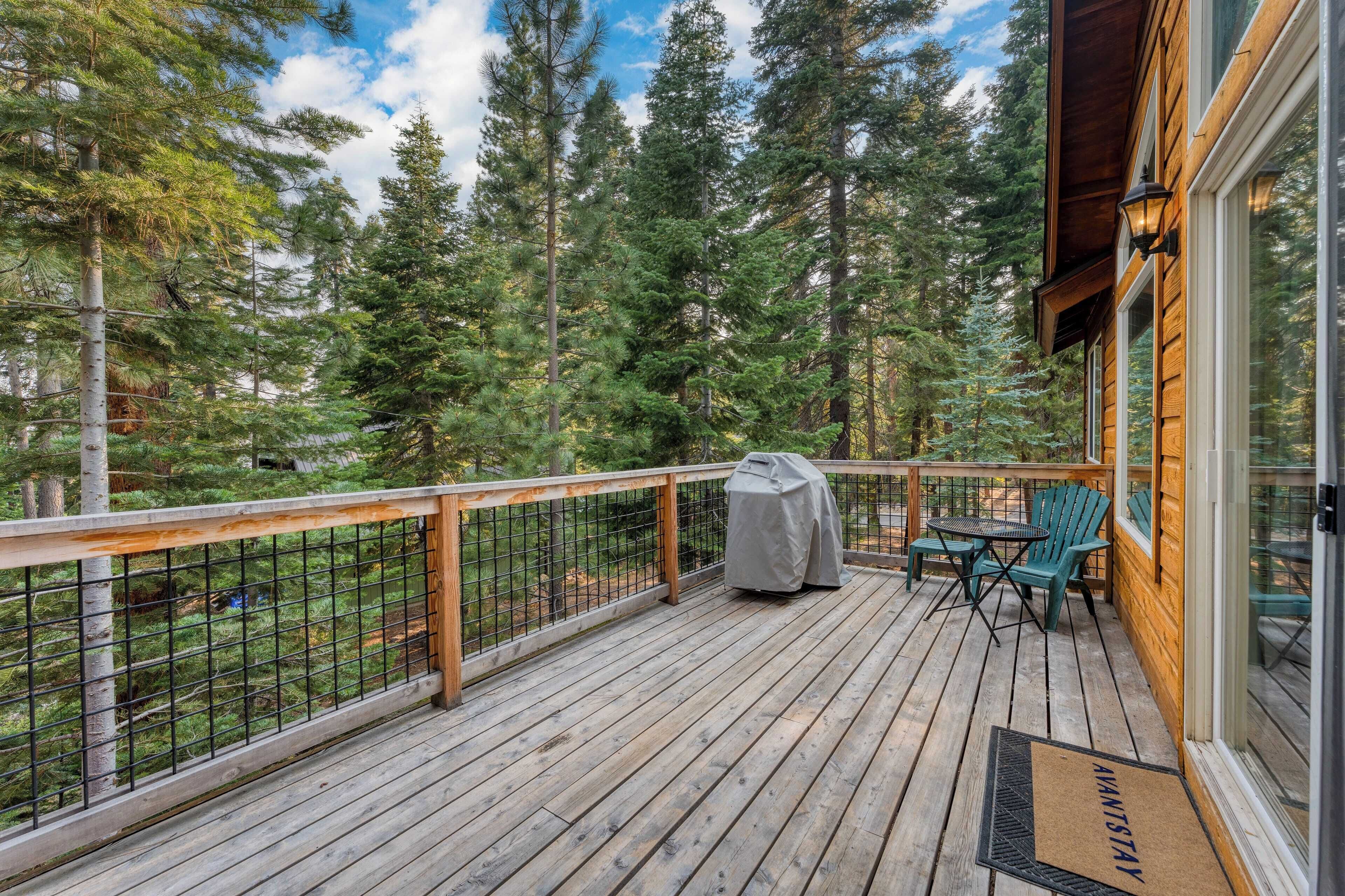 Una terraza de madera con una parrilla cubierta, asientos exteriores y vistas a un bosque.