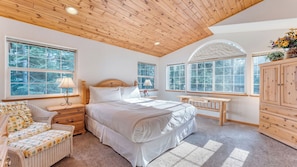 Un dormitorio con una cama grande, dos mesitas de noche, un tocador, una silla, un aparador y una ventana con vista a árboles.