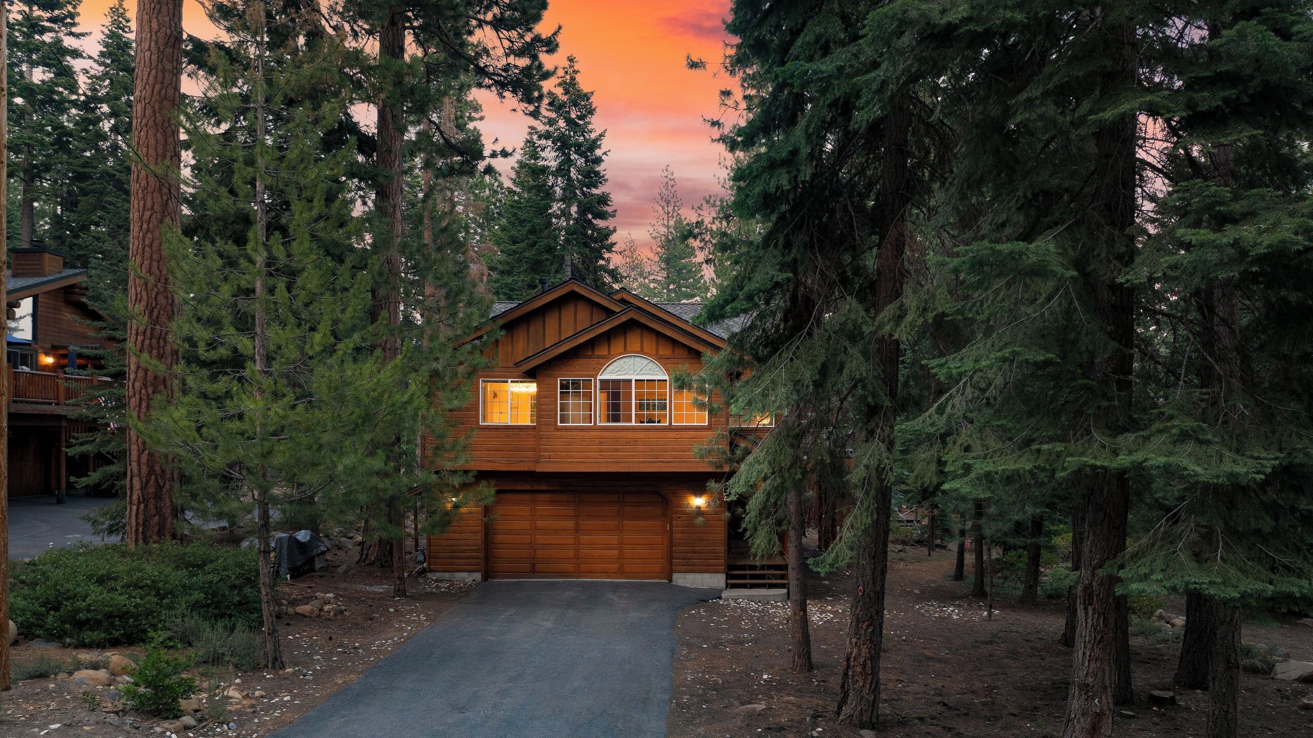 A cabin with a garage, surrounded by tall trees, at dusk.