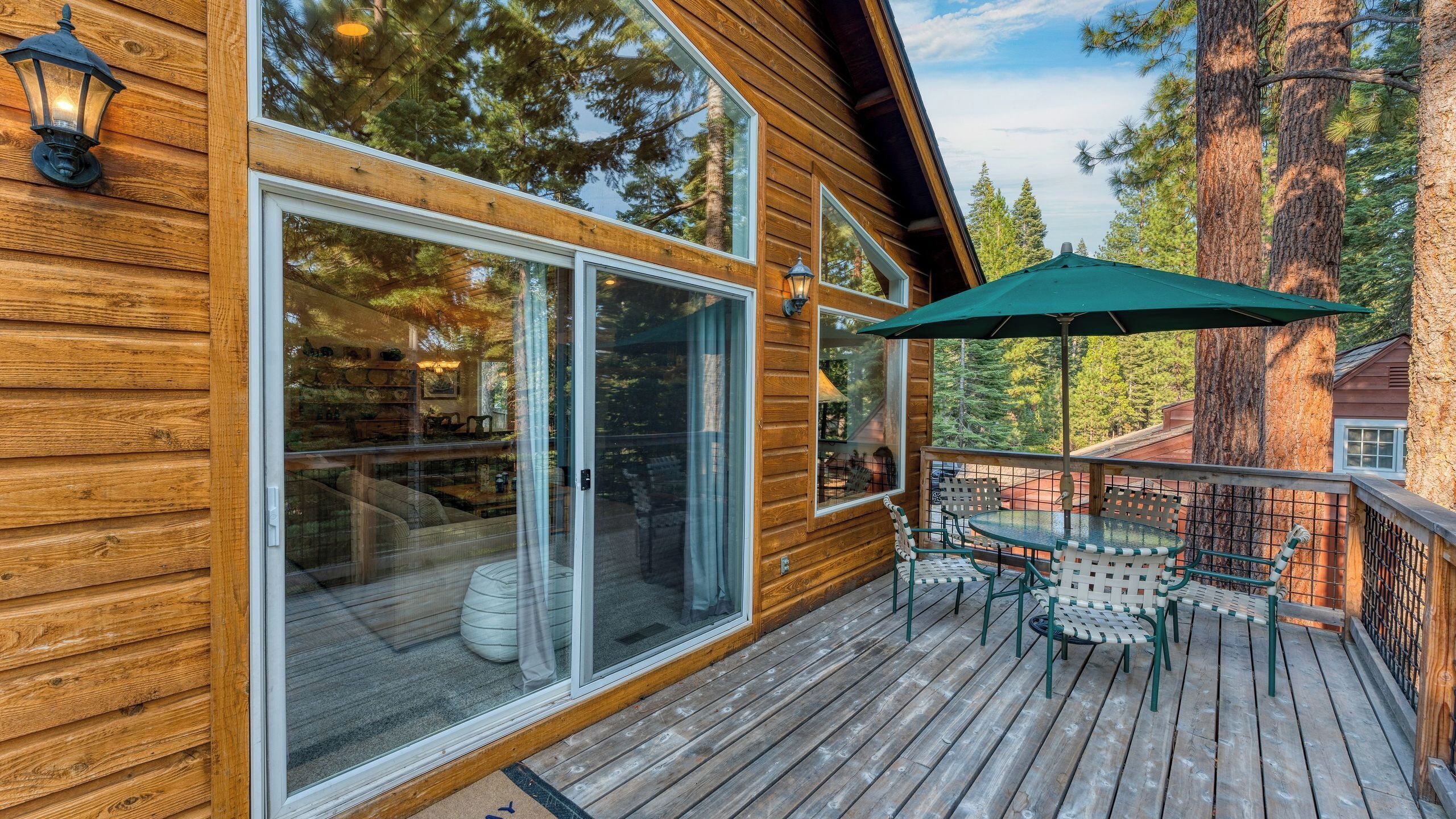 Una cabaña de madera con ventanas grandes, una terraza con mobiliario exterior y vistas a los árboles circundantes.