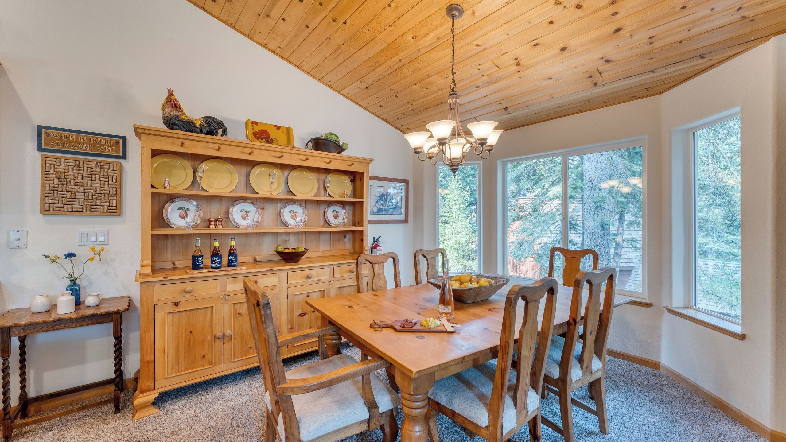 Une salle à manger en bois, avec une table, des chaises et un lustre. On y trouve des étagères remplies d’assiettes et un buffet en bois contenant des bouteilles.