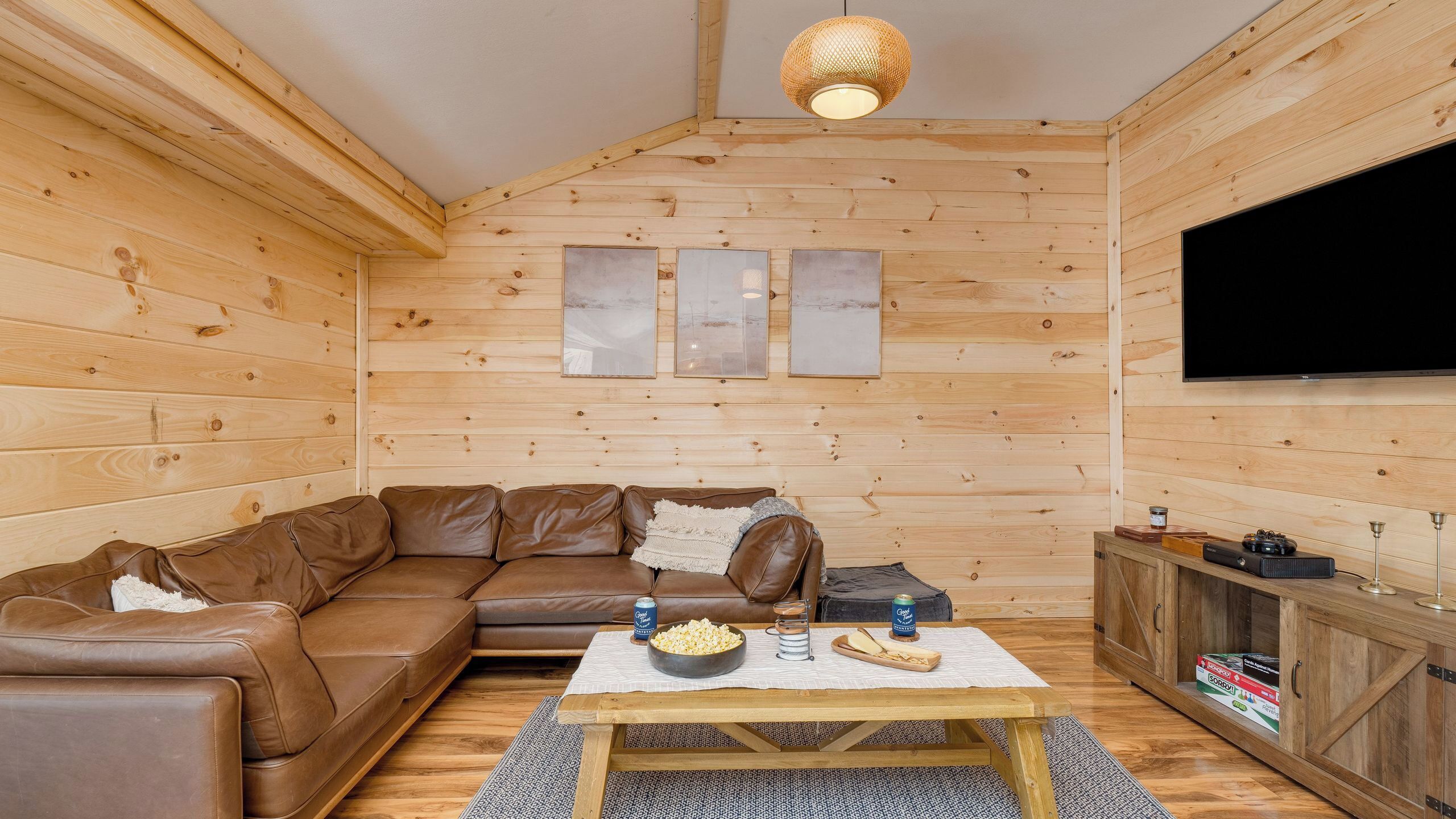 A cozy wooden interior with a brown leather sofa, a coffee table with snacks, and a flat-screen TV mounted on the wall.