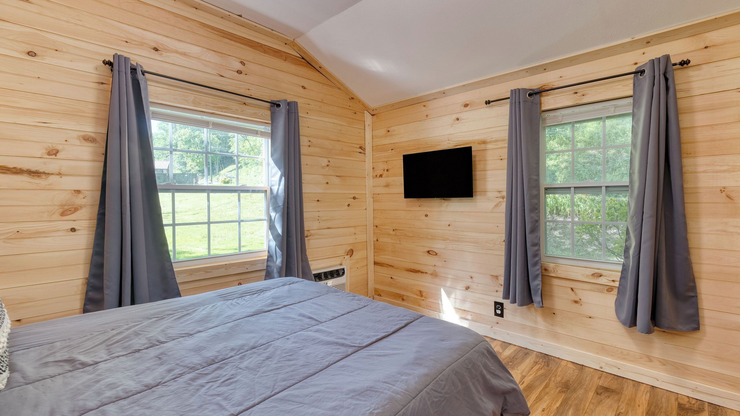 Una habitación con paneles de madera, una cama, dos ventanas con cortinas, un televisor montado en la pared y un termostato.