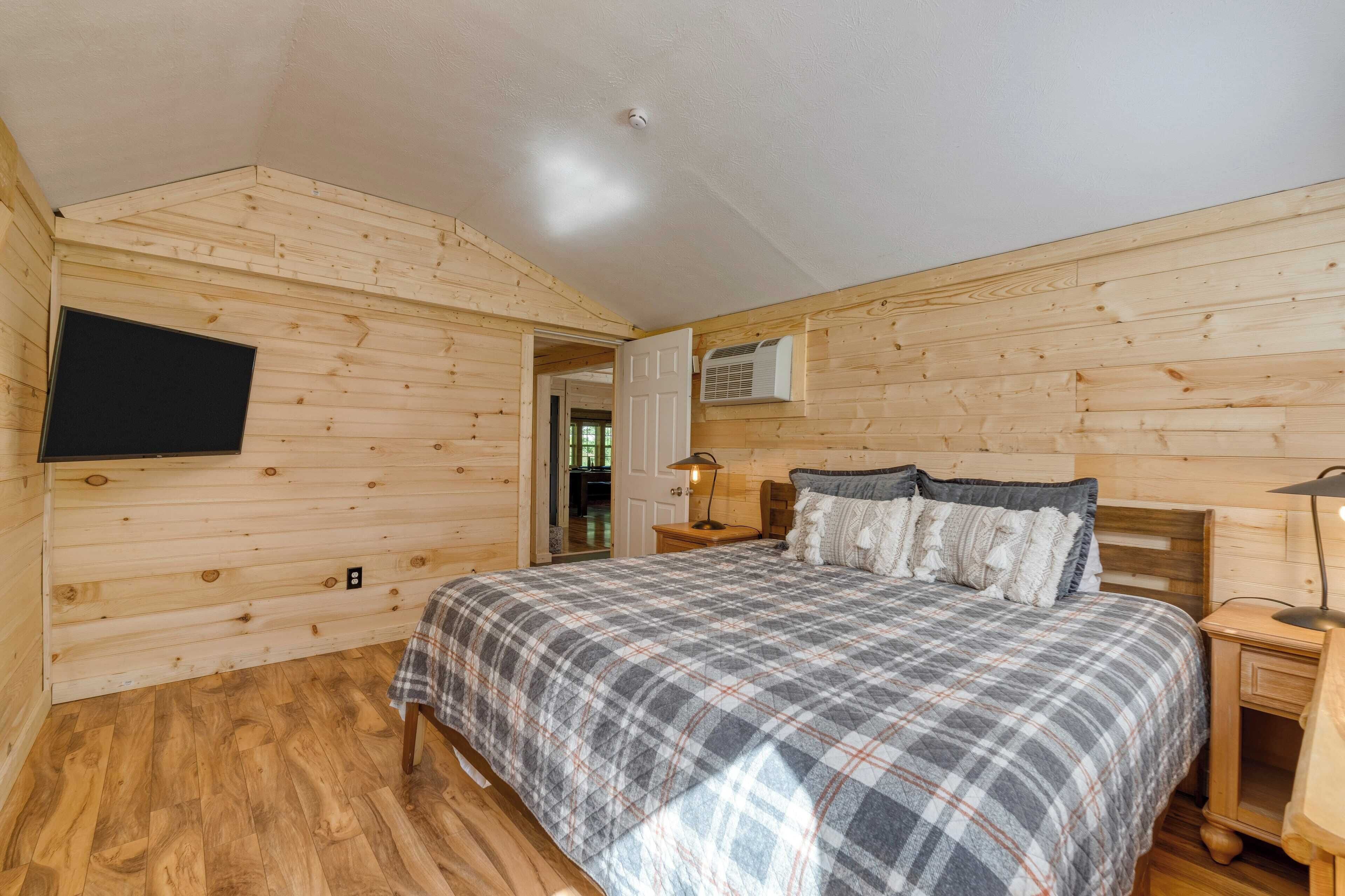 Un dormitorio con cama, paredes de madera, televisor montado y una puerta que da a otra habitación.