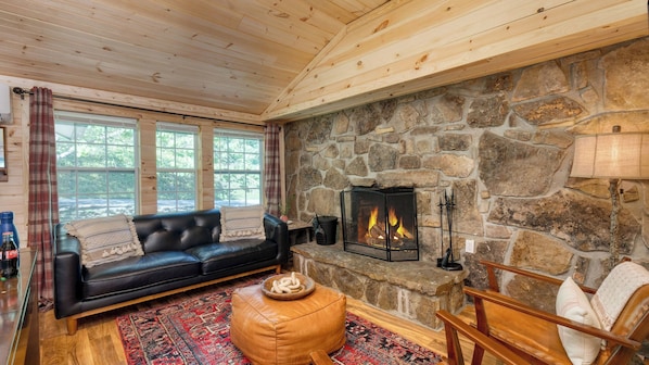 A cozy room with a stone fireplace, a black leather sofa, a wooden coffee table, and a stone wall.
