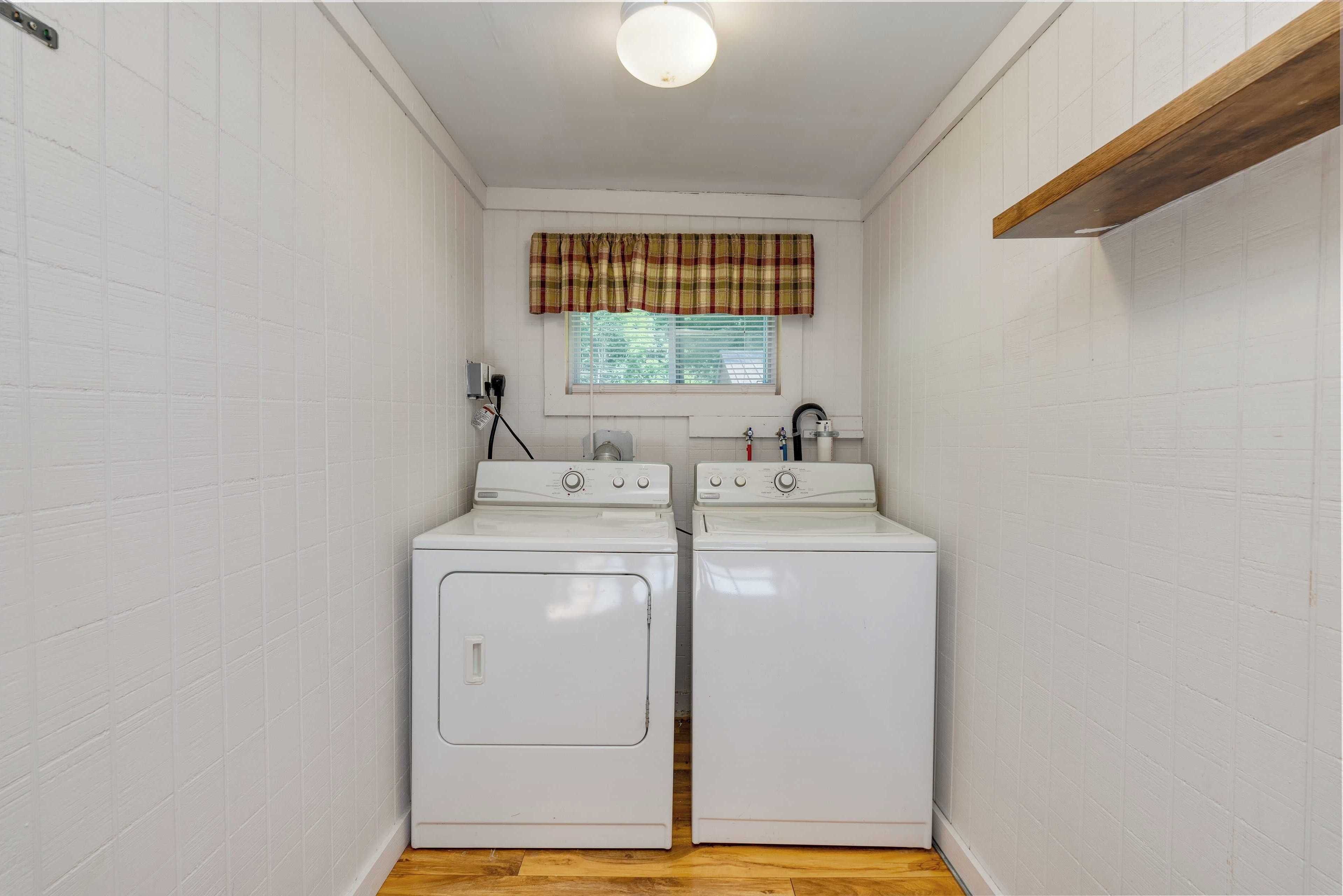A laundry room with two white washing machines, a window with a plaid valance, and a wooden shelf above the machines.