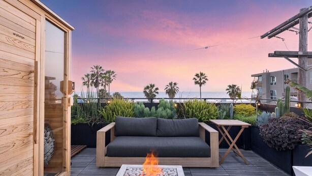 A wooden deck with a sofa and a fire feature, overlooking a sunset view with palm trees and a building in the background. - Venice View by Avantstay Walk to Venice Beach Rooftop, Ice Bath, Luxe Design (Venice)