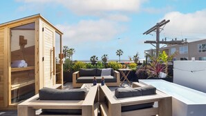 A wooden outdoor seating area with a sofa and benches, a glass-enclosed sauna, and a view of the ocean. - Venice View by Avantstay Walk to Venice Beach Rooftop, Ice Bath, Luxe Design (Venice)