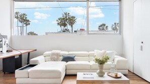 A modern living room with a large window, a white sofa, a coffee table, and a desk with a computer. - Venice View by Avantstay Walk to Venice Beach Rooftop, Ice Bath, Luxe Design (Venice)