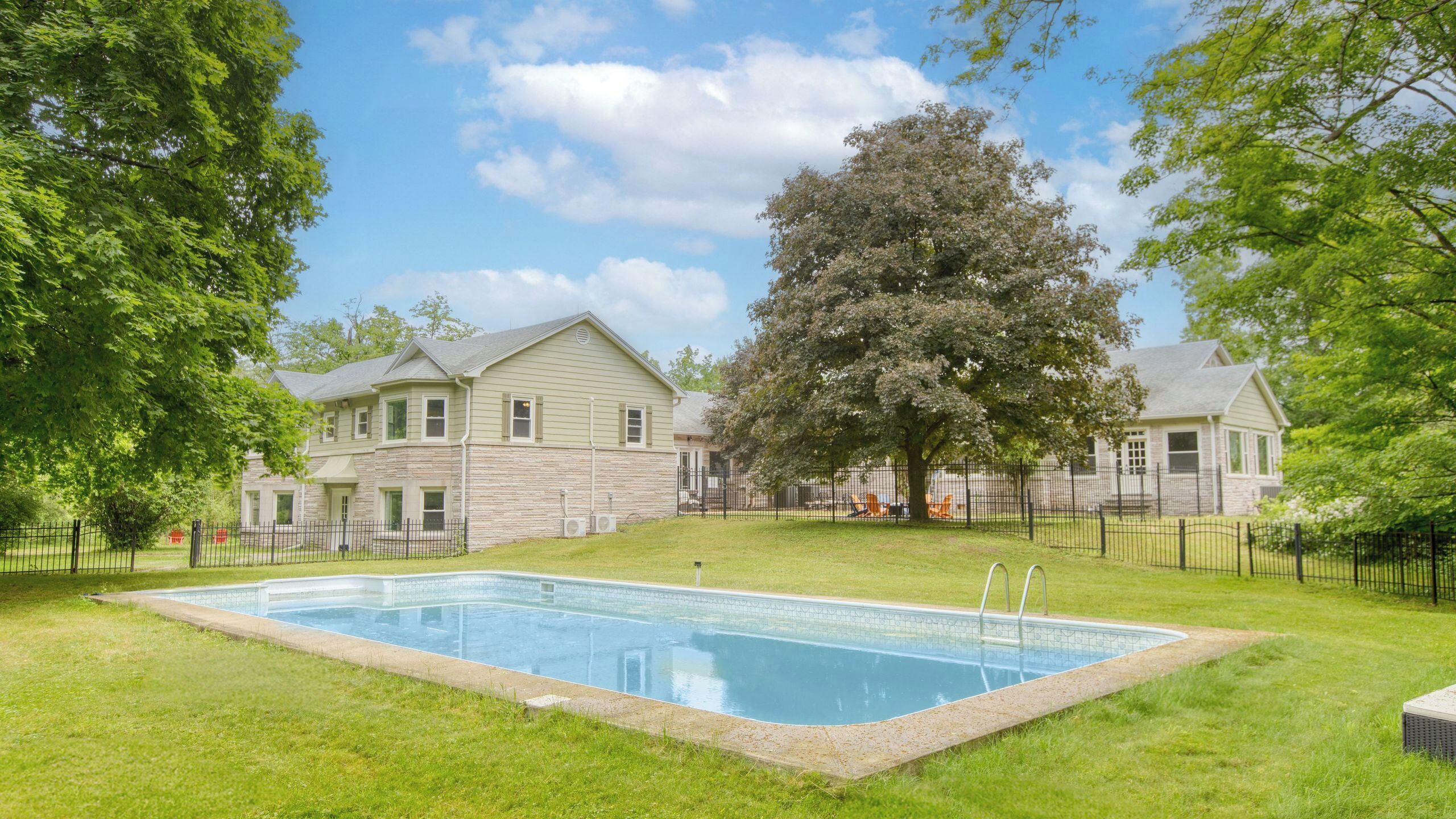 A swimming pool surrounded by a grassy lawn, with a large tree and a two-story building in the background.