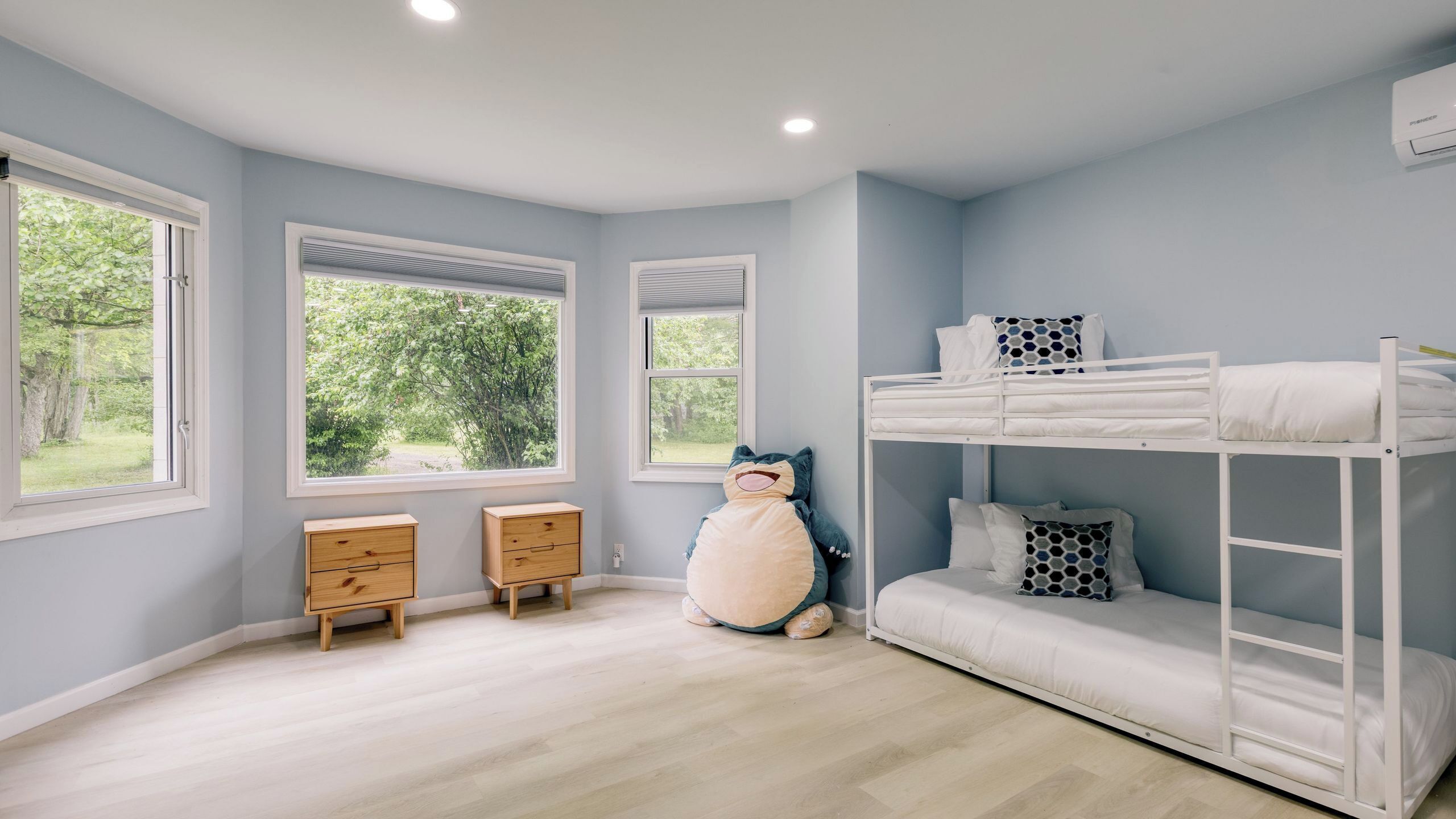 Une chambre d’enfant avec des lits superposés, deux tables de chevet en bois et de grandes fenêtres donnant sur un paysage verdoyant.