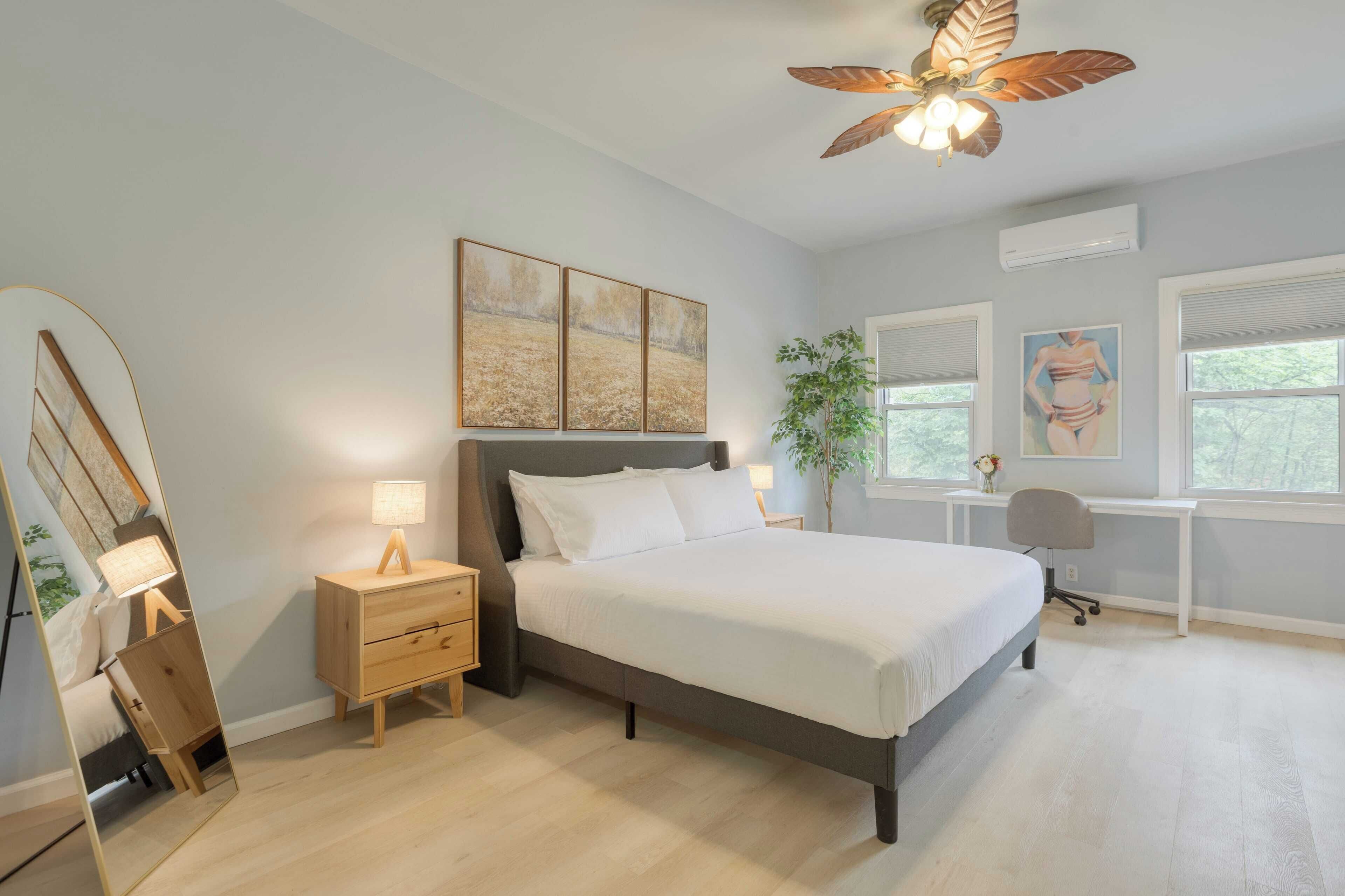 A bedroom with a bed, bedside table, lamp, desk, chair, and ceiling fan.