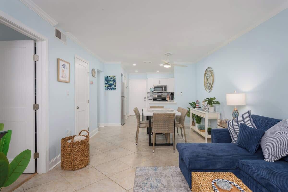 Un salon avec un canapé bleu, une table à manger avec des chaises, un coin cuisine et un couloir menant à une porte.