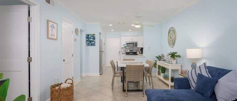 A living room with a blue sofa, a dining table with chairs, a kitchen area, and a hallway leading to a door.