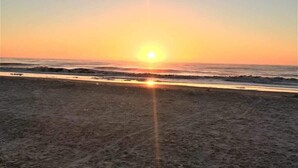 A beach at sunset with the sun low on the horizon, waves approaching the shore, and a clear sky.