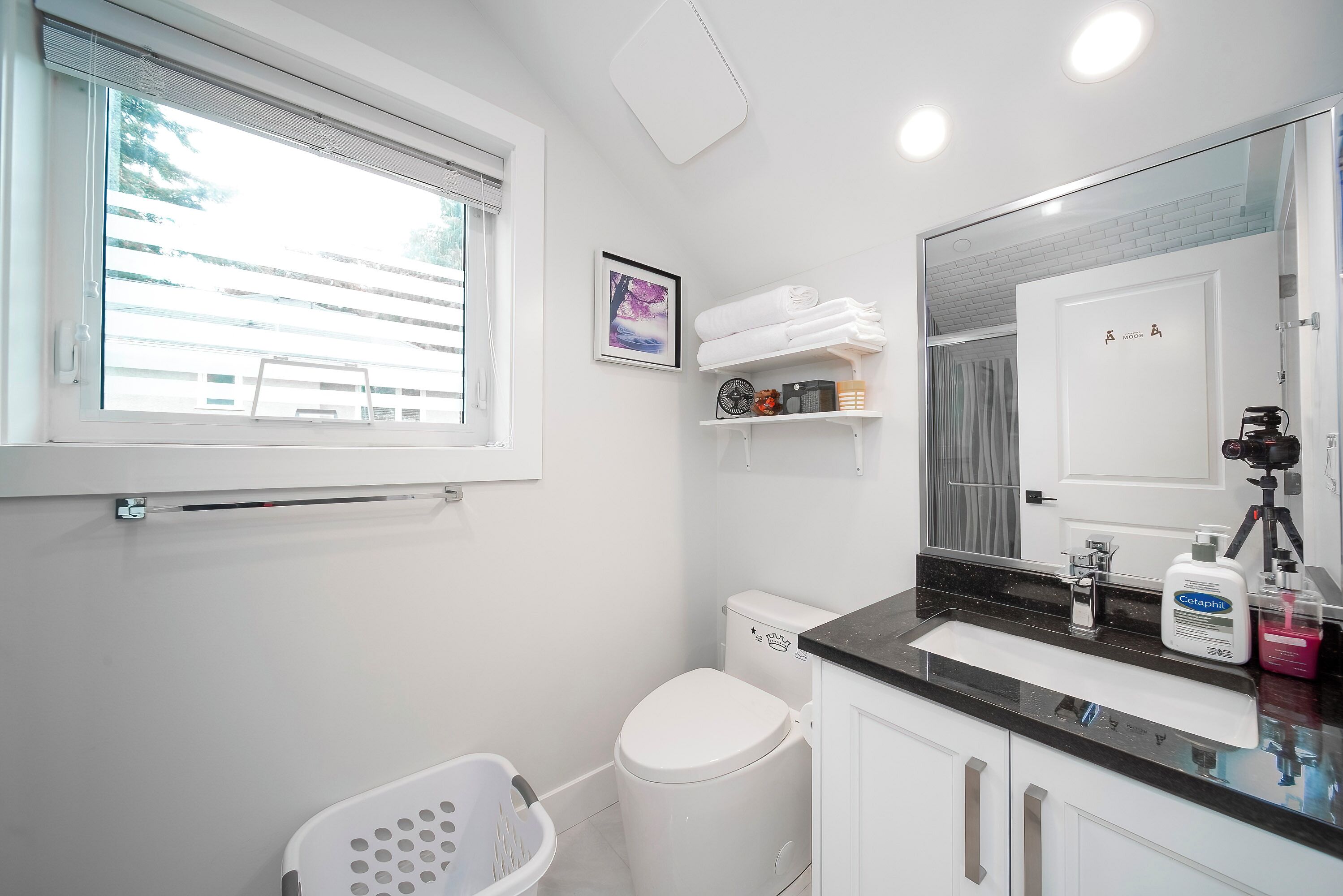 A modern bathroom with a white sink, a toilet, a window with blinds, a framed picture on the wall, and a shelf with toiletries.