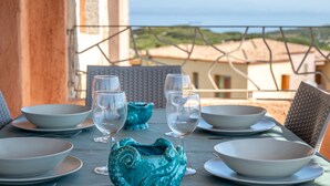 Una mesa dispuesta con cuencos blancos y copas de vino, un florero turquesa, y vista al mar y edificios en el fondo.