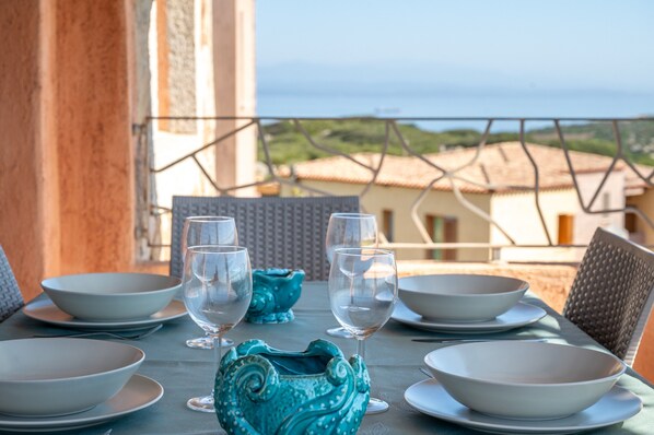 A table set with white bowls and wine glasses, a turquoise vase, and a view of the sea and buildings in the background.