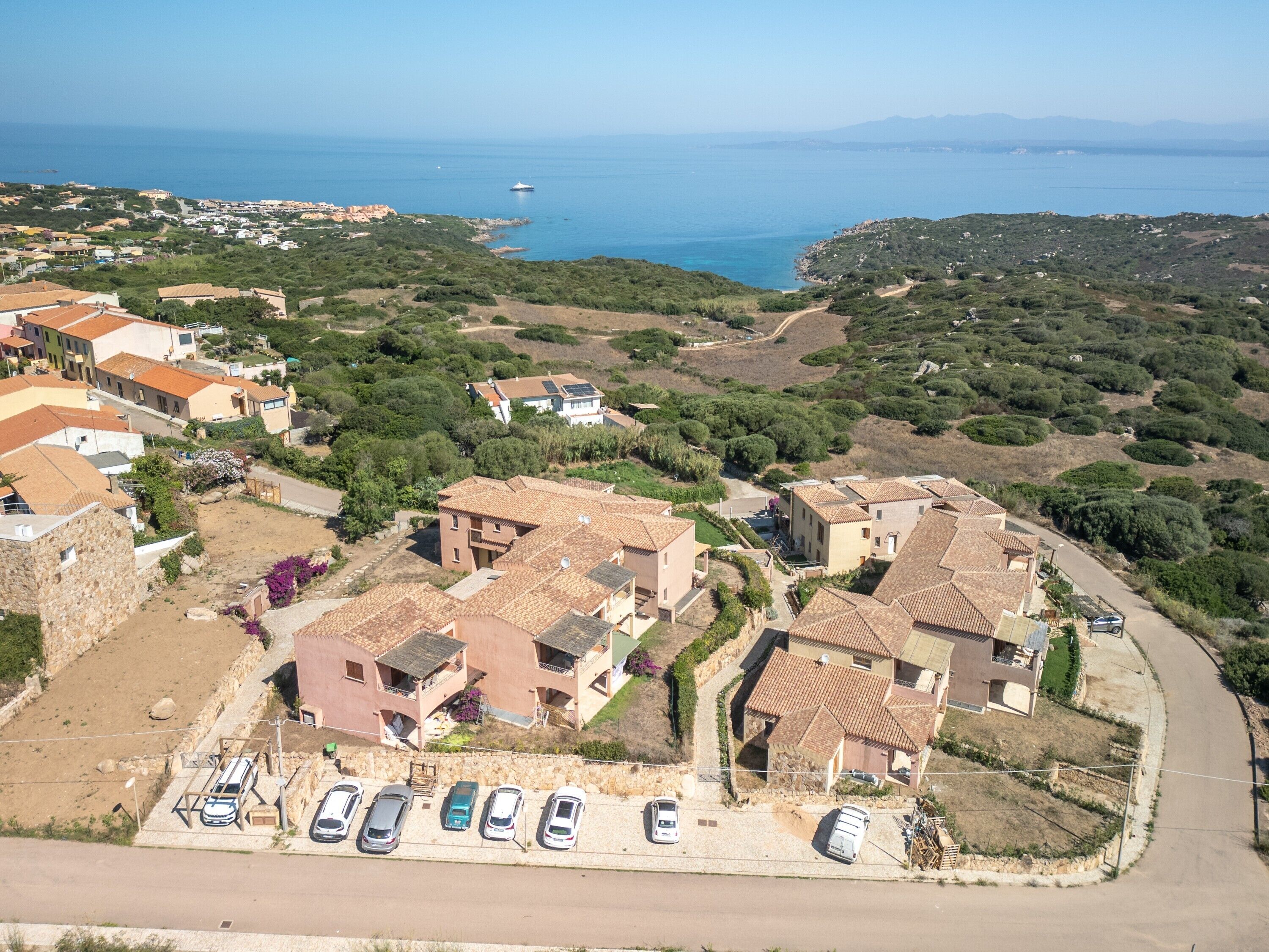 A coastal residential area with terracotta-roofed houses, a parking lot with several cars, and a view of the sea and distant mountains.