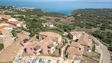 A coastal residential area with terracotta-roofed houses, a parking lot with several cars, and a view of the sea and distant mountains.