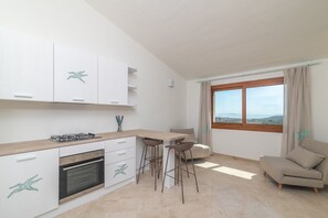 A modern kitchen with a gas stove, oven, and white cabinets. There are two bar stools and a beige sofa near a window with a view.