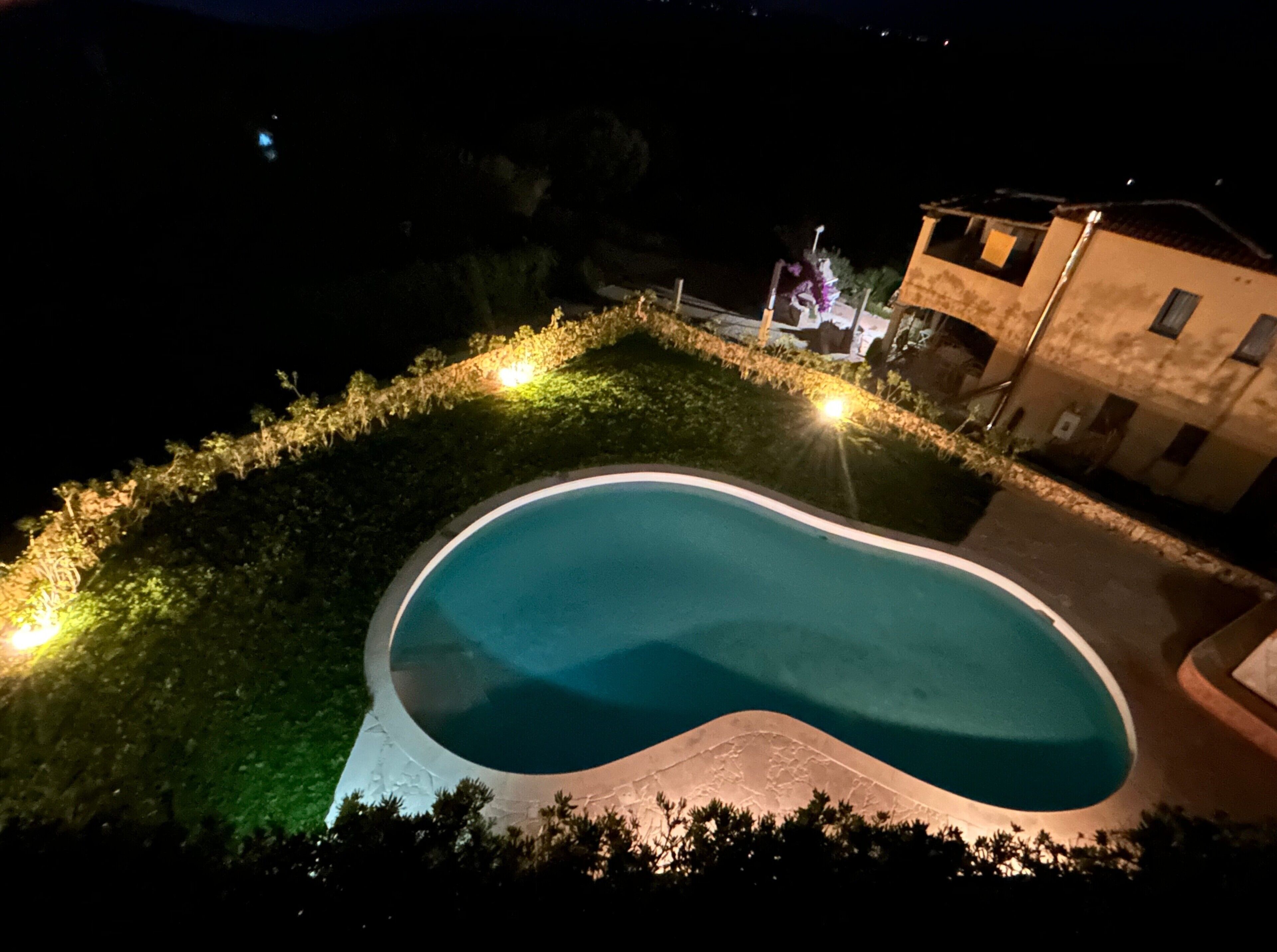 Vista notturna di una piscina a forma curva, circondata da un'area verde illuminata con luci soffuse.