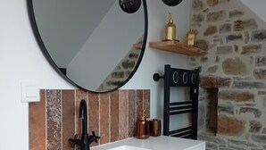 Un baño con lavamanos blanco, grifo negro y espejo redondo. Se observan dos lámparas colgantes y una pared de piedra al fondo.