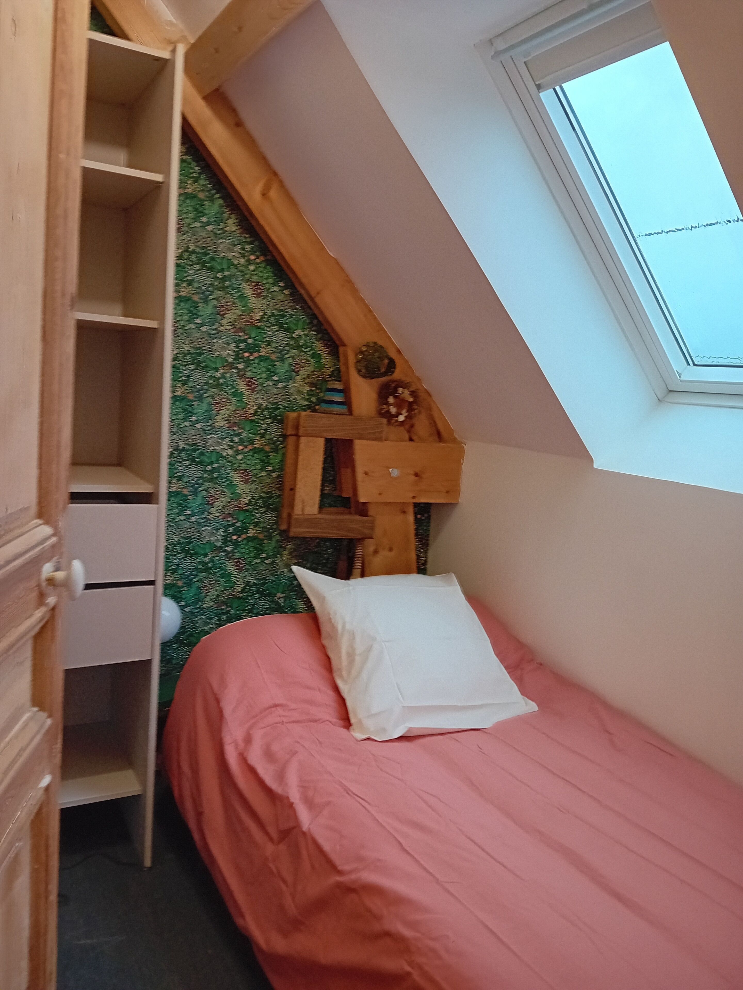 A bedroom with a sloped ceiling, a skylight, a bed with pink bedding, a wooden nightstand, and a wall with a floral pattern.