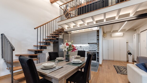 Una cucina moderna con isola centrale, un'area pranzo con tavolo e sedie in legno, e una scala che conduce a un livello superiore.