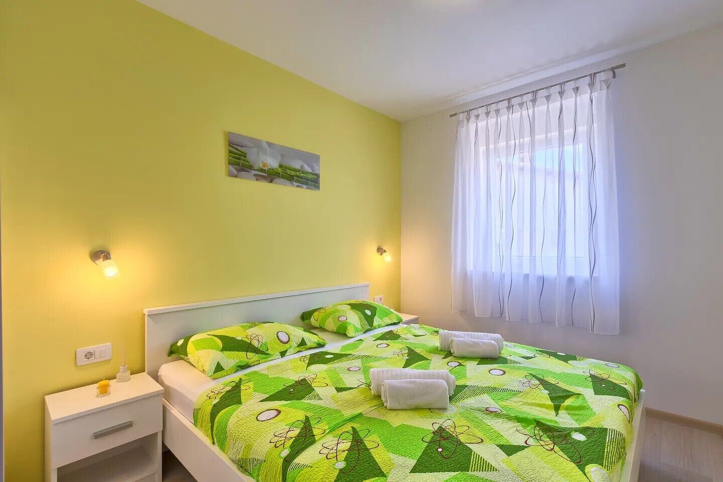 A bedroom with a green patterned bedspread, white curtains, and a small painting on the wall.