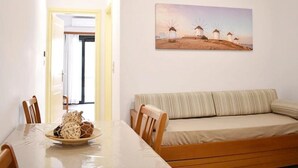 A dining area with a table, chairs, and a plate of food. There is a sofa and a painting of windmills on the wall. - Apartment 'Manolika Studio 5' with Sea View, Wi-Fi and Air Conditioning (Kythnos)