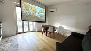 A living room with a wooden floor, a dark-colored sofa, a dining table with four chairs, and a large screen displaying a nature scene. - Suite&Chic Apartment (Bari)