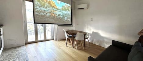 Ein Wohnzimmer mit Holzboden, einem dunkelgrauen Sofa, einem Esstisch mit vier Stühlen und einem großen Bildschirm, der eine Naturlandschaft zeigt.