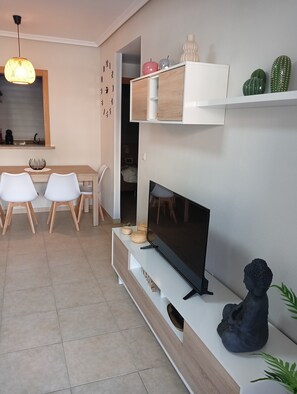 A modern living room with a flat-screen TV, a dining area with white chairs, a kitchen with a hanging light, and decorative items on shelves.