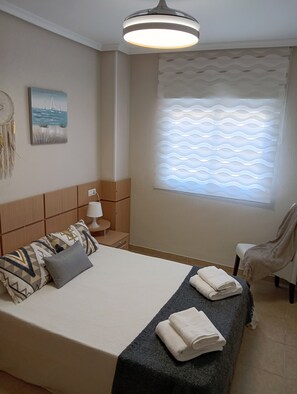 A bedroom with a bed, a nightstand, a lamp, a window with patterned blinds, and a wall with a dreamcatcher and a painting.