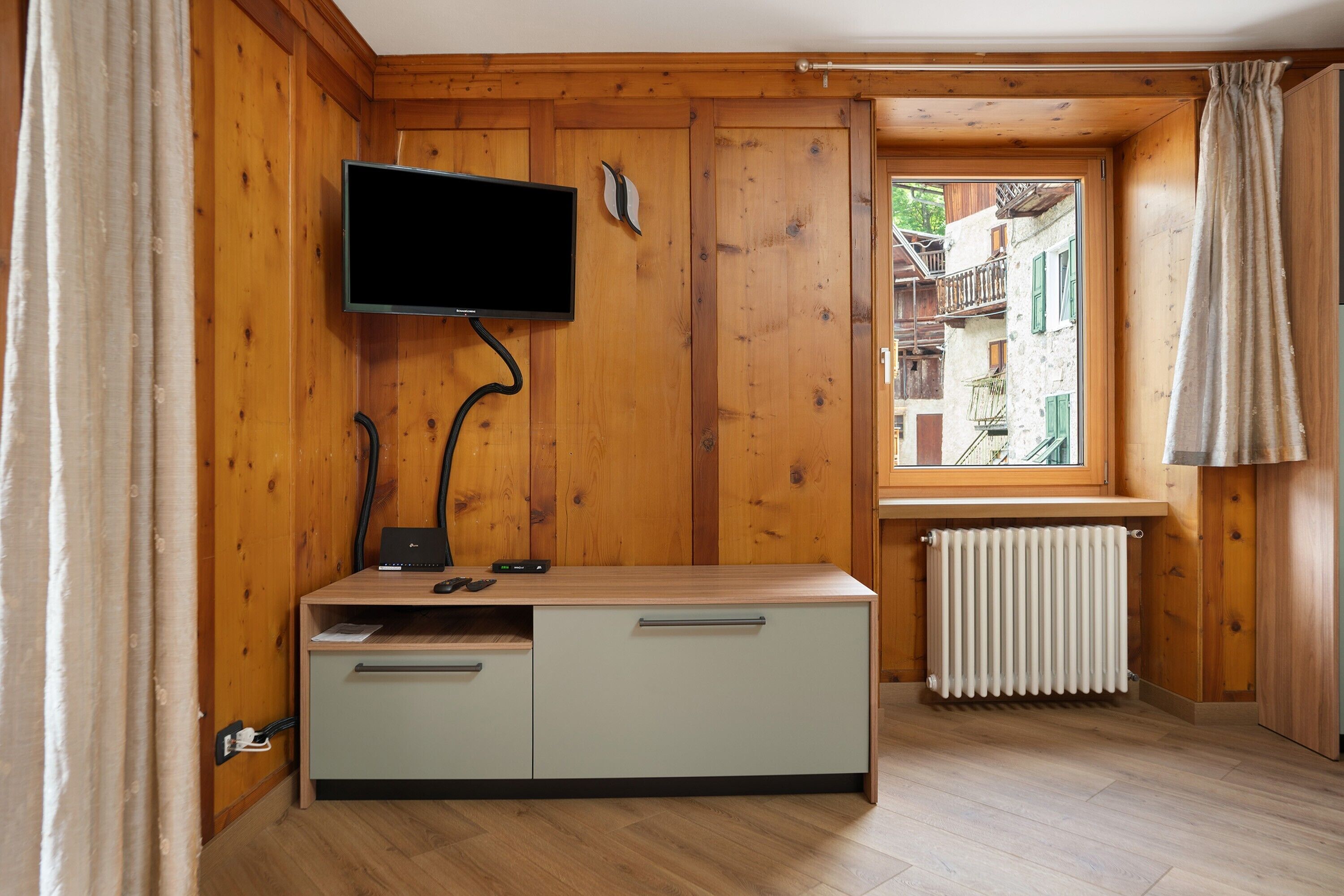 A room with wooden walls, a TV mounted on the wall, a desk with drawers, a radiator, and a window with a view of buildings outside.