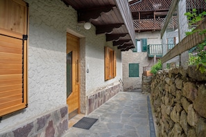 Une cour dallée de pierres, avec des volets en bois, un mur de pierre et une porte en bois.