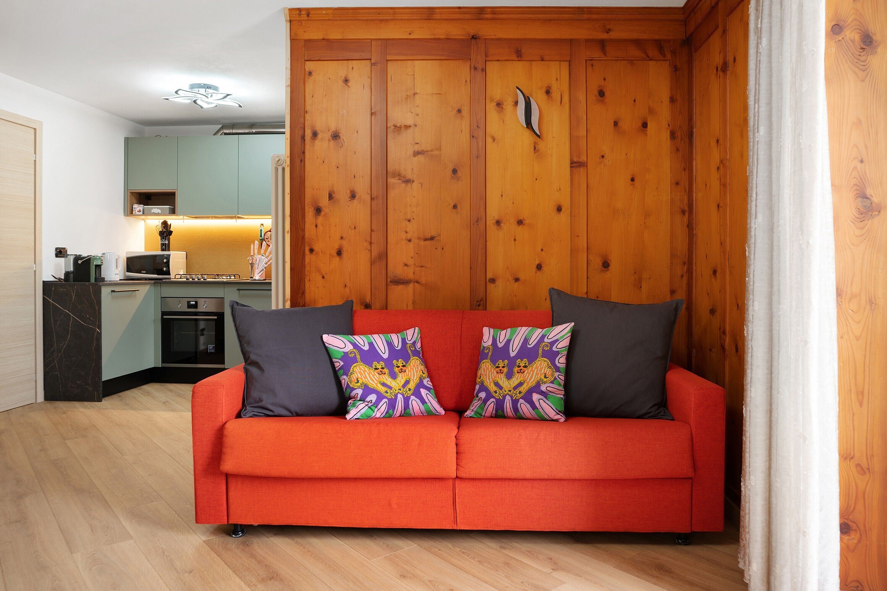 A red sofa with patterned cushions in a room with wooden walls and a kitchen in the background.
