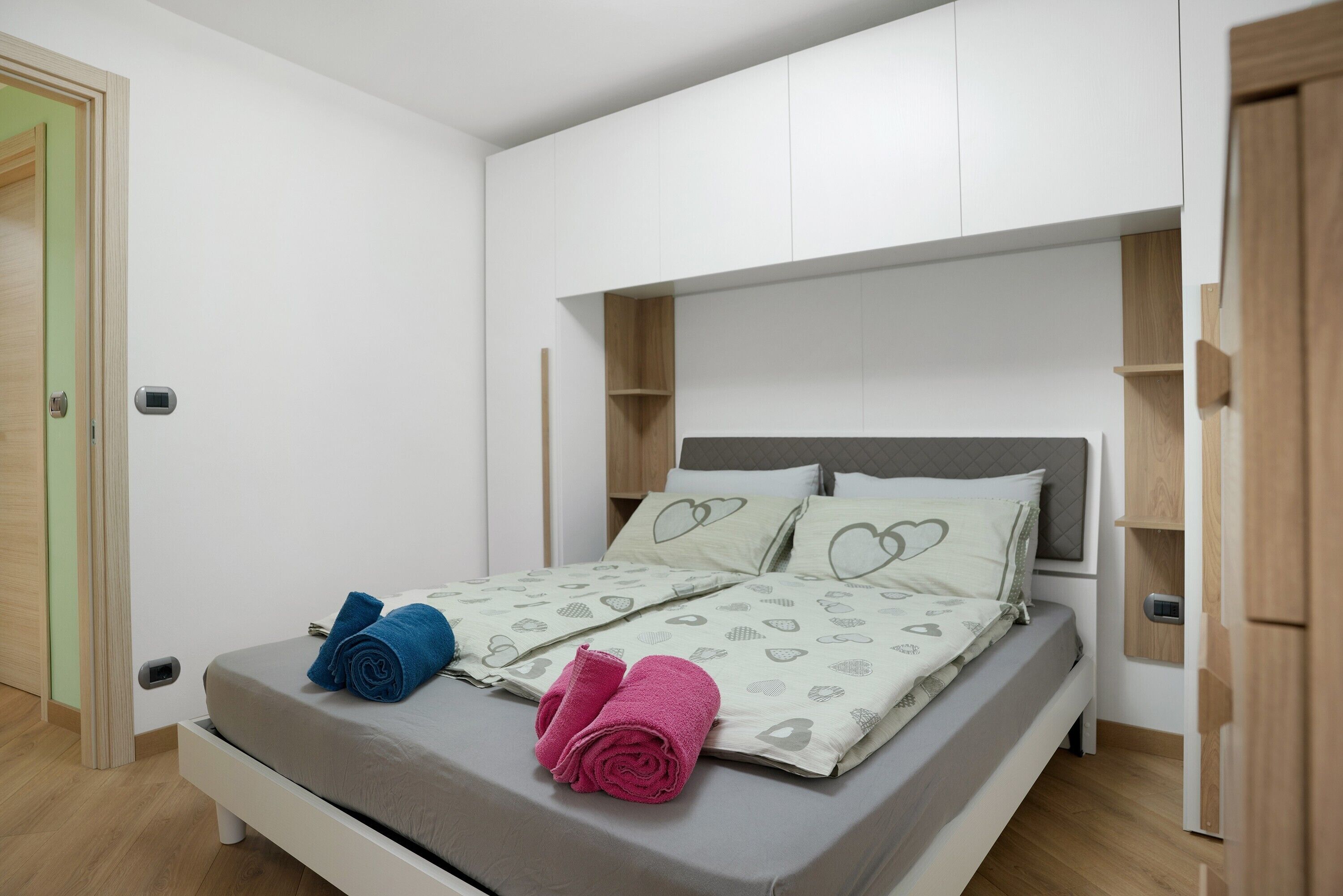 Un lit bien fait, avec des serviettes pliées, une tête de lit dotée d’étagères intégrées et un mur de couleur claire.