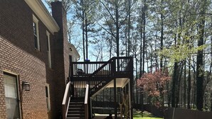 A residential backyard with a brick house, a wooden deck, and a staircase leading to a second-story balcony.