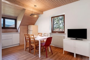 A dining area with a white table, wooden chairs, a red chair, a ceiling light, a window, a radiator, and a TV on a white cabinet.