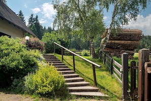 A stone staircase leading to a building with a tiled roof, surrounded by lush greenery and a stack of firewood.